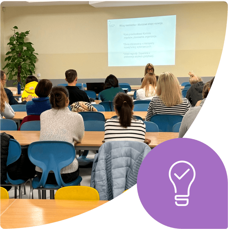 Grupa osób słucha wykładu w sali, na ekranie wyświetlana jest prezentacja.