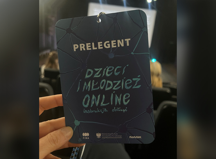 Osoba trzyma identyfikator prelegenta konferencji "Dzieci i młodzież online" na tle sali.