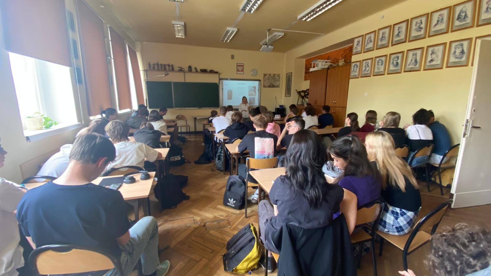 Uczniowie siedzą w klasie i słuchają nauczycielki prowadzącej lekcję przy tablicy multimedialnej.