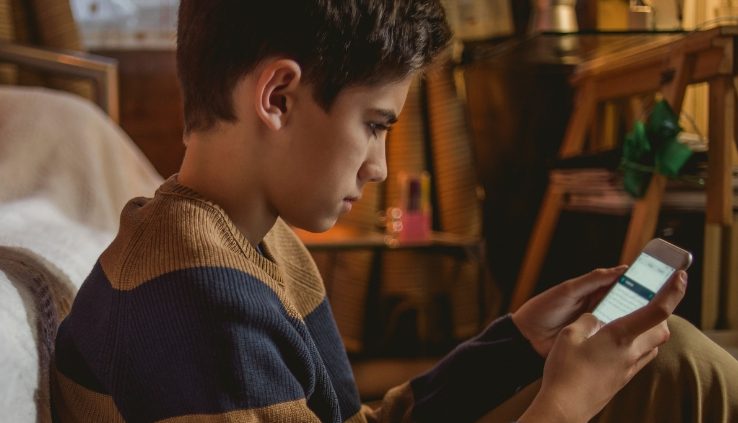 Chłopiec siedzi na kanapie i patrzy na ekran smartfona w dłoniach.